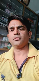 Portrait of a man in a yellow shirt indoors, seated with shelves in the background.