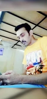 Man in a yellow shirt relaxing with phone under a bed.