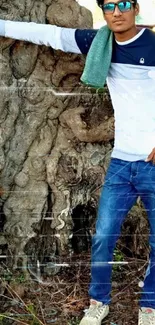 Man casually posing against a large tree trunk with blue sunglasses.