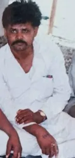 Man in white outfit seated outdoors in a casual setting.