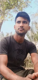 Man sitting outdoors amidst greenery, wearing a casual black shirt.