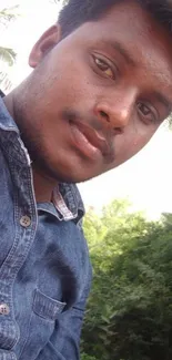 Young man in denim shirt outdoors with greenery.