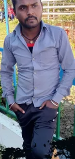 A man in a blue shirt posing outdoors by a fence.