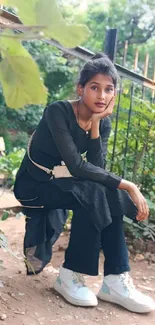 Person in black attire sitting outdoors, surrounded by greenery.