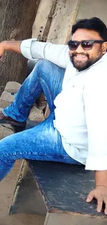 Man in jeans and sunglasses sitting casually outdoors.