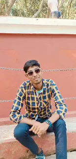 Young man in checkered shirt sits against an orange wall.