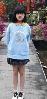 Girl in blue sweater stands on garden bridge with flowers.