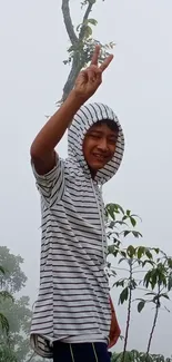 Young man in hoodie making peace sign in misty nature.