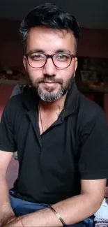 Casual portrait of a bearded man in a room, wearing glasses and a black shirt.