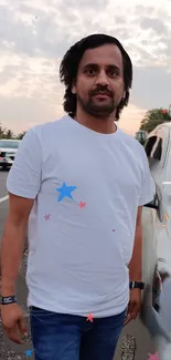 Man in white t-shirt standing by road with cars and sky.