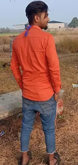 Man in orange shirt stands in rural landscape.