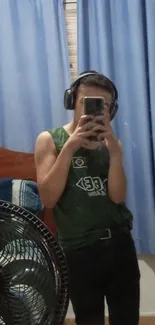 Casual selfie in a mirror with headphones, featuring blue curtains and a fan.