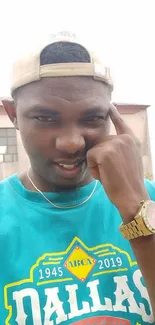 Man in teal t-shirt poses with beige cap and gold watch in urban setting.