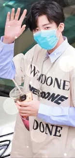 Individual in beige shirt and mask, holding a drink, waving casually.