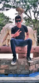 Man in sunglasses sits casually in an urban park with trees and statues.