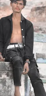 Young man in casual urban attire sitting against a stone background.