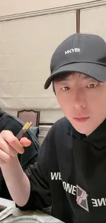 Young man dining with chopsticks, wearing a casual cap and hoodie.