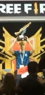 Gamer holds trophy on stage with Free Fire logo backdrop.