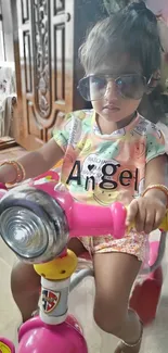 Child with sunglasses on pink bike indoors.