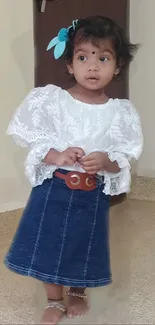 Cute toddler in denim skirt with white lace top, standing indoors.