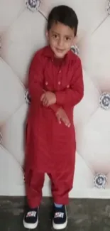 Smiling child in red traditional attire standing against a decorative wall.