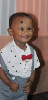 Smiling child in suspenders with a red bow tie against a soft background.