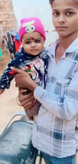 Boy holding a baby outdoors with brick wall background.