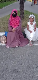 Two people sitting outdoors, a woman in pink and a girl in white, on a serene street.