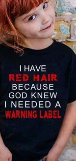 Redhead child in a shirt with funny text against a brick wall.