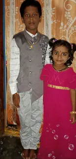 Sister in pink dress and brother standing indoors, smiling.