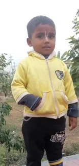 Toddler in yellow hoodie outdoors with greenery background.