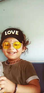 A smiling child with a cap and sunglasses, holding a bottle.