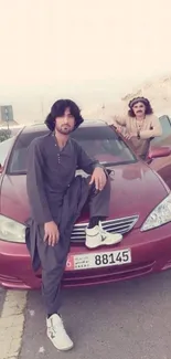 Two people posing on a red car on a scenic road.