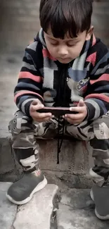 Young child in winter attire focused on a smartphone, sitting on a brick surface.