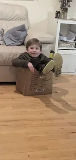 Child seated in a cardboard box near a cozy beige couch.