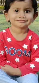 Smiling child in red star shirt sitting on grass.