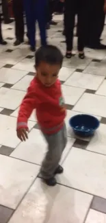 Child in a red sweater on a tiled floor with blue bowl nearby.