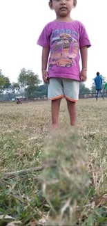 Child standing in grassy field with a purple shirt, enjoying a sunny day.