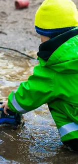 Child in green jacket playing with a toy truck in a puddle.