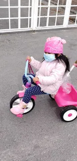 Child on a pink tricycle with a bright hat and mask on a sunny day.