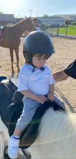 Young child riding a pony on a sunny day outdoors, wearing a safety helmet.