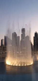 Fountain illuminated against city skyline at dusk.