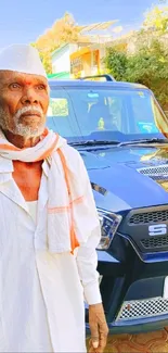 Elderly man in traditional attire beside a classic blue car.