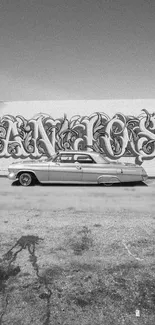 Black and white image of a classic car in front of graffiti on a wall.