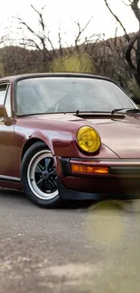 Classic maroon Porsche with yellow headlights on a scenic road.