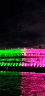 A dam lit with vibrant green, pink, and purple lights at night reflecting on water.