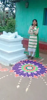 A woman stands beside vibrant Rangoli art in front of a teal house.