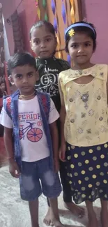 Three children in colorful outfits standing indoors, smiling at the camera.