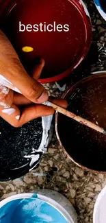 Hand holding a brush over colorful paint cans, vibrant wallpaper.