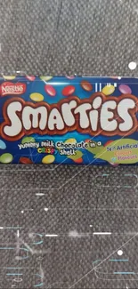 Smarties candy box on gray fabric background, showcasing colorful candies.